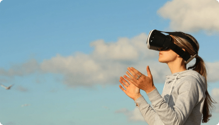 A person with long hair wearing a virtual reality headset stands against a backdrop of a clear blue sky with scattered clouds.