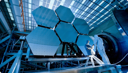 An engineer in a clean suit inspects a large, hexagonal mirror segment of a giant telescope in an industrial setting. The intricate structure of the telescope and the scale of the engineering project are emphasized by the surrounding scaffolding and blue-tinged lighting